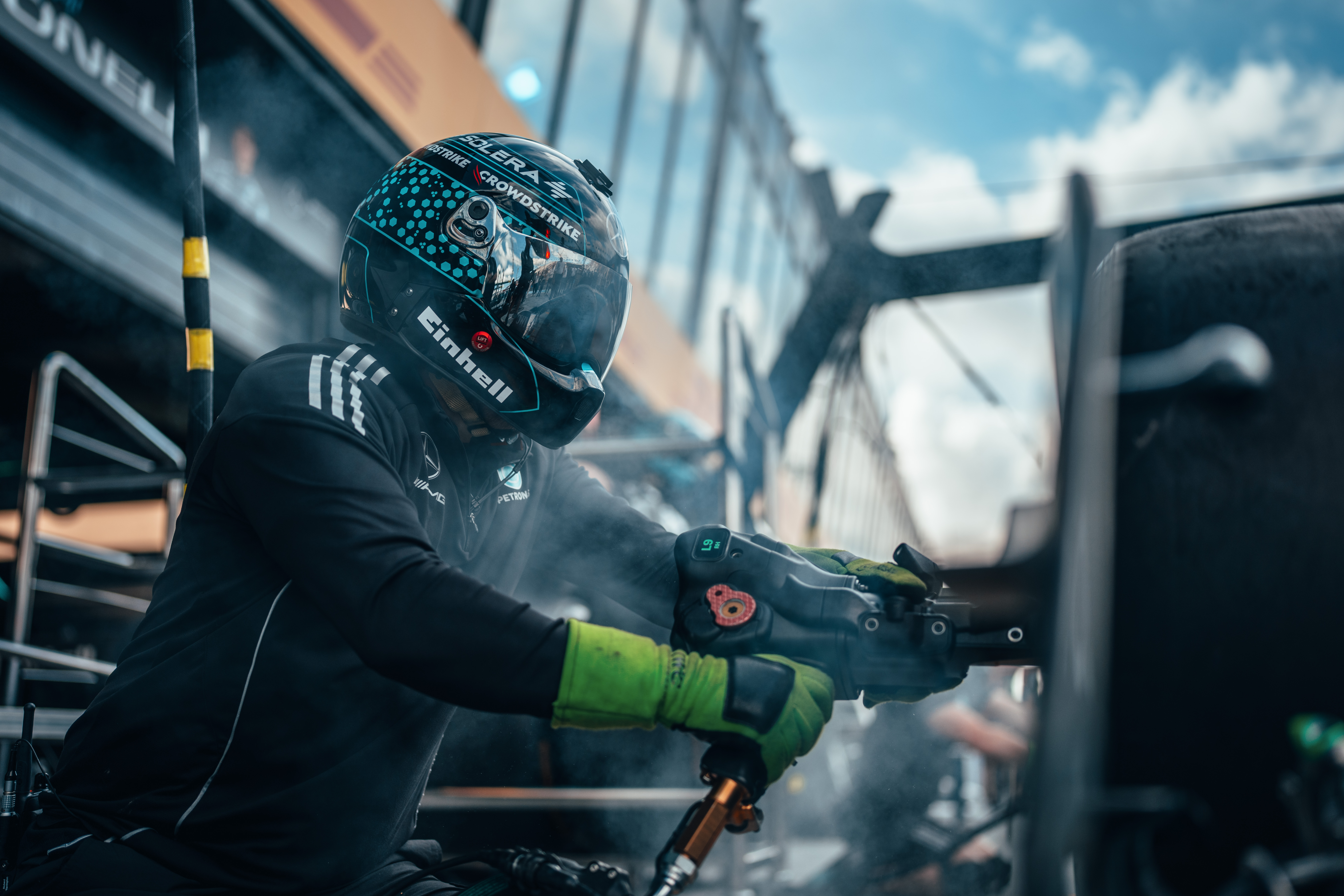 A Formula 1 pit crew member wearing a helmet and gloves uses a pneumatic wheel gun during a pit stop, with smoke and equipment around.